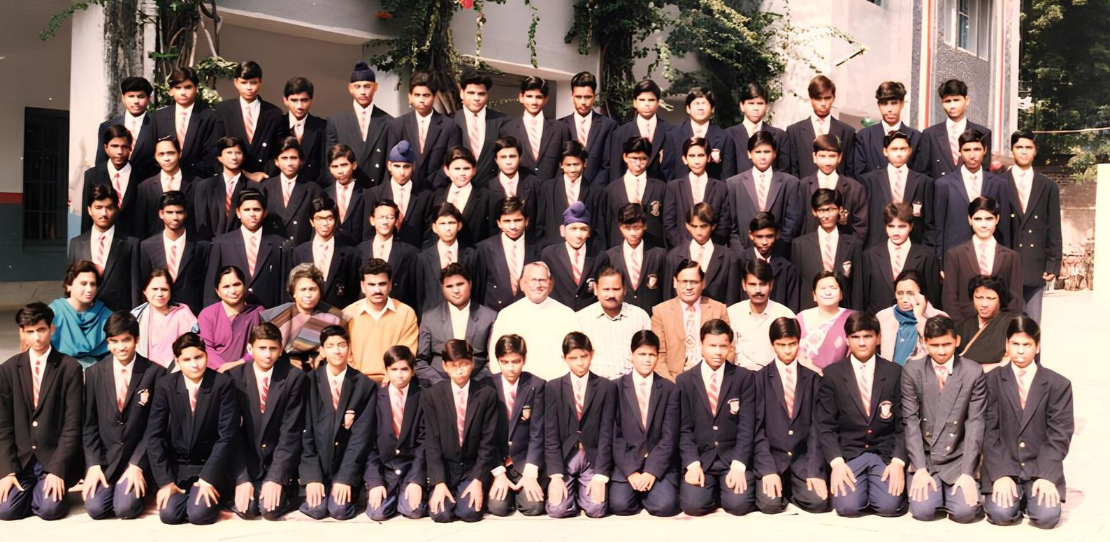 School group photo of students and faculty in formal attire with blazers and ties, taken outside a school building with greenery and flowers in the background, featuring multiple rows of students and teachers from [specific year or school if known].