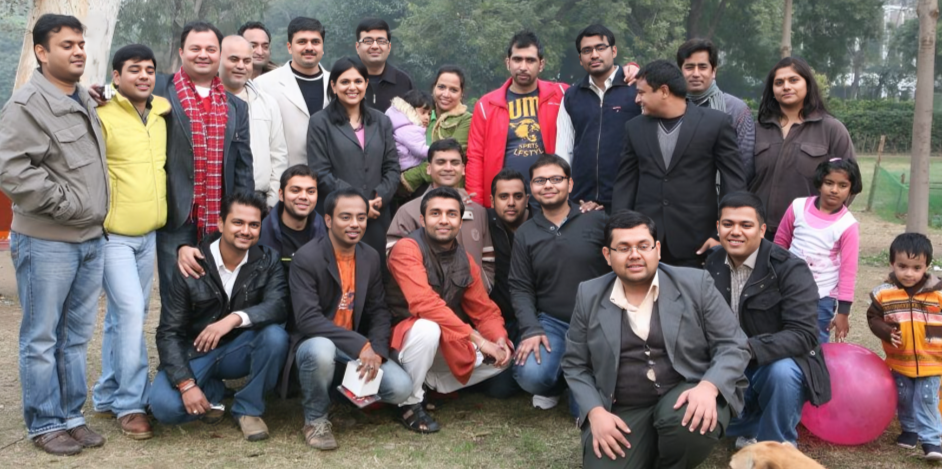 Outdoor group photo of friends and family gathered in a park, smiling and dressed in winter attire, with green trees and grass in the background, celebrating togetherness and friendship in a natural setting